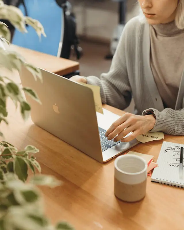 BLOG IMAGE - Jeffrey Studios (2) A person sitting at a wooden desk uses a laptop, with one hand on the keyboard and the other holding a sticky note. A notebook, pen, and coffee mug are also on the desk; a plant is partially visible in the foreground.